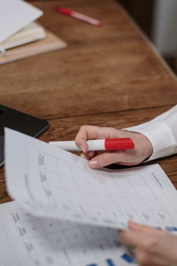 Close-up shot of a hand with a red marker examining charts on paper on a wooden desk.